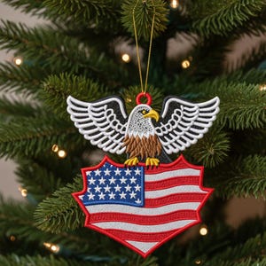 May include: Embroidered Christmas tree decoration featuring an American bald eagle with outstretched wings above a shield displaying the U.S. flag. The ornament is red, white, blue, black, and brown, with a gold hanging cord. The decoration is on a green Christmas tree.