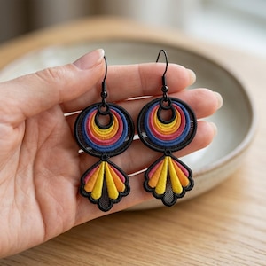 May include: A pair of dangle earrings featuring black embroidered circular and fan-shaped motifs. The design includes concentric rings of yellow, orange, pink, and blue thread on a black mesh background, suspended from black metal earring hooks.