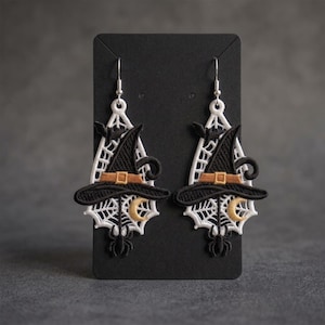 May include: A pair of dangling earrings featuring embroidered white spiderwebs, black witch hats with orange bands, small black bats, gold crescent moons, and black spiders hanging from silver-toned metal hooks on a black display card.