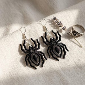 May include: A pair of black spider earrings with silver-colored hooks. The spider earrings are made of a detailed, embroidered material. A ring with a black stone and a sprig of lavender are also in the image.