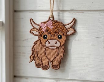 Könnte beinhalten: Gestickter Ornament einer Highland-Kuh mit rosa Schleife. Die Kuh ist braun mit hellbraunen Akzenten und großen, dunklen Augen. Aufgehängt an einer natürlichen Schnur. Das Ornament ist mit detaillierten Stichen gestaltet.