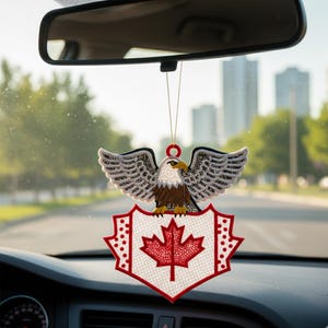 Puede incluir: Un ambientador para coche con un diseño de águila bordada, con las alas extendidas, sobre un escudo que muestra una hoja de arce roja sobre un fondo blanco. El ambientador está colgado del espejo retrovisor de un coche.