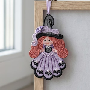May include: Embroidered Halloween witch ornament. The witch has red curly hair, a black hat, and a purple dress with pearl accents. The ornament is hanging from a lavender ribbon and is framed by a wooden border.