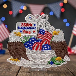 Pode incluir: Decoração patriótica bordada com duas águias emoldurando uma cesta cheia de elementos vermelhos, brancos e azuis. A cesta contém uma pequena bandeira americana, bolas listradas e esferas estreladas. Uma placa diz "250th Independence Day."