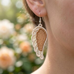 May include: A close-up of a single earring shaped like a wing. The wing is made of cream-colored lace with a brown center. The earring is suspended from a silver hook. The background is blurred with soft green and orange tones.