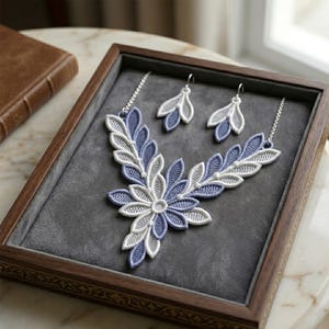 Può includere: Un delicato set di collana e orecchini presentato in una scatola di legno. I gioielli presentano un design floreale con ricami bianchi e blu. Il set è posto su uno sfondo di velluto grigio.