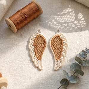 May include: Two embroidered angel wing patches with a light beige and brown color scheme. The wings have a detailed, textured design and a small silver loop at the top for hanging. A spool of brown thread sits nearby.