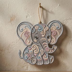 K&ouml;nnte beinhalten: Gestickter Elefanten-Ornament in Grau, mit pastellfarbenen rosa, blauen und cremefarbenen Wirbeln. Der Elefant hat gro&szlig;e Ohren und einen R&uuml;ssel und h&auml;ngt an einer Juteschnur. Das Ornament ist mit einem goldenen Haken an einer Wand befestigt.