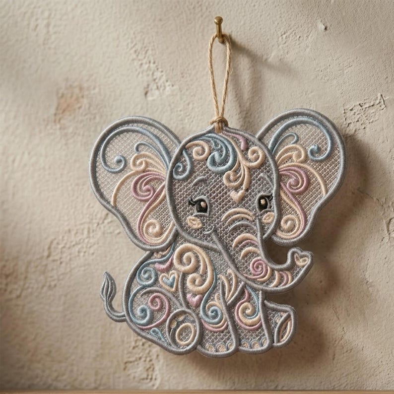K&ouml;nnte beinhalten: Gestickter Elefanten-Ornament in Grau, mit pastellfarbenen rosa, blauen und cremefarbenen Wirbeln. Der Elefant hat gro&szlig;e Ohren und einen R&uuml;ssel und h&auml;ngt an einer Juteschnur. Das Ornament ist mit einem goldenen Haken an einer Wand befestigt.