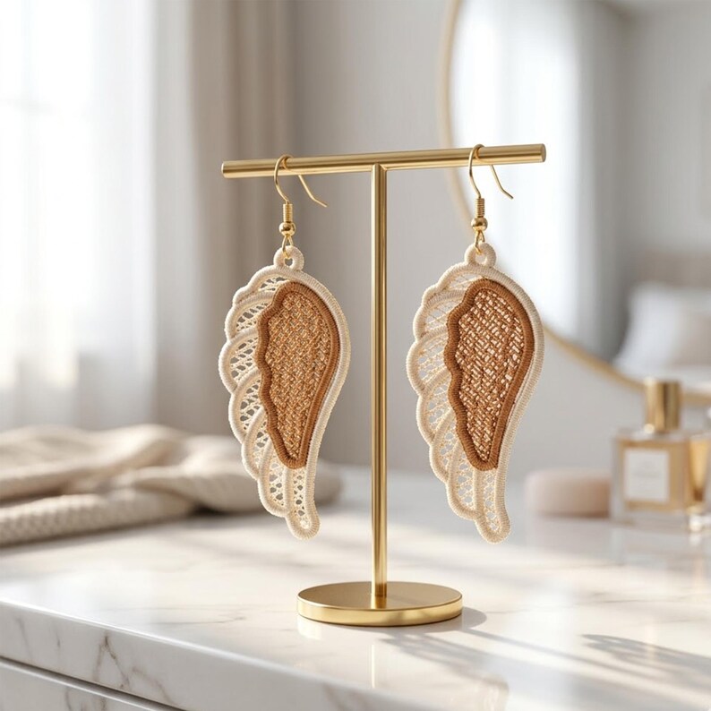 May include: A pair of gold-toned dangle earrings shaped like angel wings. Each earring features a beige, lace-like outer wing and a textured, brown inner wing. The earrings hang from gold-colored hooks and are displayed on a gold earring stand.