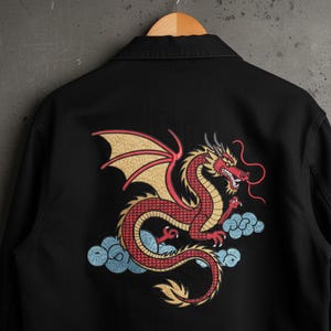 May include: Black denim jacket featuring a detailed embroidered dragon design on the back. The dragon is red with gold wings and accents, set against blue cloud motifs. The jacket is hanging on a wooden hanger.