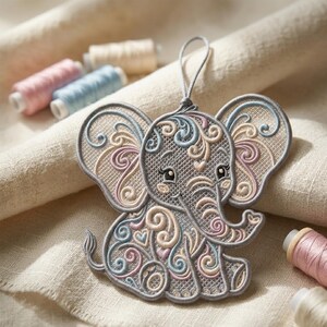 K&ouml;nnte beinhalten: Gestickter Elefanten-Ornament in Grau mit pastellfarbenen rosa, blauen und beigen Wirbeln und Herzdetails. Der Elefant hat eine graue Umrandung und eine Schlaufe zum Aufh&auml;ngen. Garnrollen in passenden Farben befinden sich im Hintergrund.