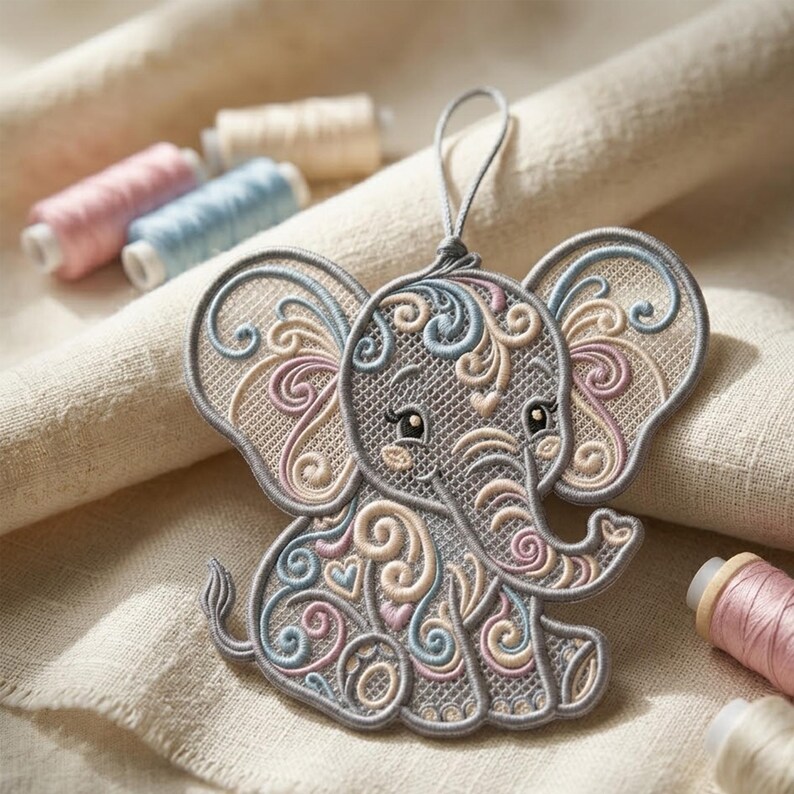 K&ouml;nnte beinhalten: Gestickter Elefanten-Ornament in Grau mit pastellfarbenen rosa, blauen und beigen Wirbeln und Herzdetails. Der Elefant hat eine graue Umrandung und eine Schlaufe zum Aufh&auml;ngen. Garnrollen in passenden Farben befinden sich im Hintergrund.