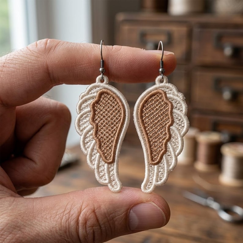 May include: A pair of angel wing earrings. Each earring features a detailed, embroidered wing design with a cream-colored outer edge and a tan-colored center. The earrings are held up by silver-colored hooks.