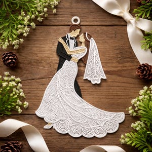 Könnte beinhalten: Ein weiß-schwarzes, gesticktes Hochzeitsornament, das ein Brautpaar in Umarmung zeigt. Das Brautkleid und der Schleier der Braut sind aus detaillierter Spitze. Der Bräutigam trägt einen schwarzen Smoking. Das Ornament befindet sich auf einem Holzgrund mit Grün und Band.
