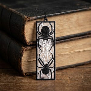 May include: A rectangular bookmark featuring intricate white lace embroidery depicting a spider web pattern. Two black embroidered spiders are positioned vertically on the lace, one near the top and one near the bottom. A small black loop is at the top.