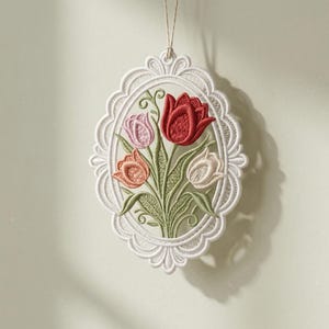 Może przedstawiać: Biała, owalna, haftowana ozdoba z bukietem tulipanów w odcieniach czerwieni, różu i brzoskwini. Kwiaty osadzone są na zielonych łodygach i liściach, obramowane delikatną, białą koronkową obwódką. Do zawieszenia przymocowany jest cienki sznurek.