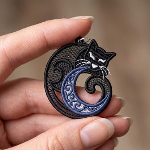 May include: Black cat pendant with a crescent moon design. The cat is embroidered in black with white whiskers, and the moon is blue with a detailed pattern. The pendant is held by a hand.