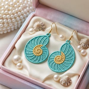 May include: A pair of turquoise and gold seashell earrings with gold-colored hooks, presented in a pink and blue box with pearls and seashells. The earrings have a detailed embroidered design, resembling a nautilus shell.