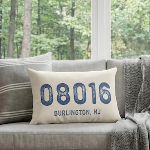 Könnte beinhalten: Ein Wohnzimmer mit einem hellgrauen Sofa, Kissen und einem Holztisch. Ein rechteckiges Kissen zeigt den Text "08016 BURLINGTON, NJ" in Blau. Eine große Topfpflanze und ein Weidenkorb ergänzen die natürliche Einrichtung.