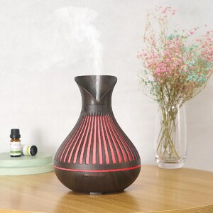 May include: A dark brown essential oil diffuser with a vase-like shape. The diffuser has vertical red lines and emits a stream of vapor. Two small essential oil bottles and a vase of flowers are in the background.