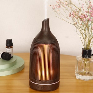 May include: A dark brown essential oil diffuser with a vertical ribbed design. The diffuser has a curved shape and a small opening at the top, with steam rising from it. The base has two buttons. A small bottle of essential oil is visible.