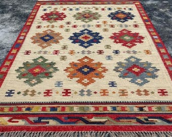 May include: A rectangular beige rug featuring a repeating floral pattern in red, blue, green, and orange. The rug has a red border with a geometric design and fringe along the edges. The rug is made of woven material.