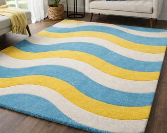 May include: A rectangular area rug with a wavy pattern in shades of blue, yellow, and white. The rug is placed on a dark wood floor, and the design creates a sense of movement and visual interest. The rug is a decorative element for a living room.