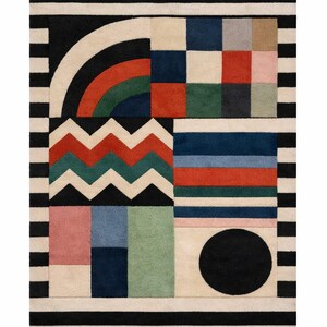 May include: Geometric patterned rug with a black and white striped border. The rug features a variety of shapes and colors, including a rainbow arch, zigzags, squares, and a black circle. The color palette includes red, green, blue, pink, and beige.