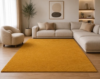 May include: A rectangular area rug in a solid mustard yellow color with a short, dense pile texture. The rug is positioned on a light-toned wooden floor, extending from the foreground toward a large sectional sofa and a matching armchair.