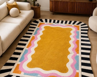 May include: A rectangular area rug with rounded corners featuring a central mustard yellow field surrounded by a wavy pink border, light blue accents, and an outer frame of alternating black and white stripes. The rug has a tufted texture.