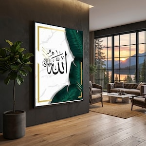 May include: Framed wall art with Arabic calligraphy in black on a white marble background, framed by a gold border. Green tropical leaves adorn the right side. The artwork is displayed in a modern living room setting with a view.