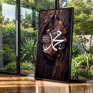 May include: Framed artwork showing a detailed brown leaf with white Arabic calligraphy. The leaf is set against a dark wood-grain background. The artwork is displayed indoors near a window with a view of greenery.