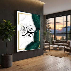 May include: A framed artwork with Arabic calligraphy in black on a white marble effect background, framed with gold and green tropical leaves. The art is displayed in a modern living room setting.