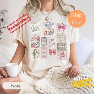 May include: Ivory-colored t-shirt featuring a collage of Mahjong-themed illustrations, including tiles, dice, and bows. The design includes the text "MAHJ" and "ONE MORE GAME". The shirt has a relaxed fit and is made of a soft material.