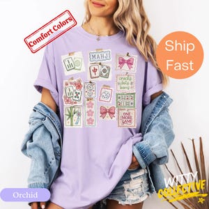 May include: Orchid-colored t-shirt featuring a collage of Mahjong-themed illustrations. The design includes tiles, dice, and decorative elements with the text "MAHJ" and "ONE MORE GAME". The shirt has short sleeves and a relaxed fit.