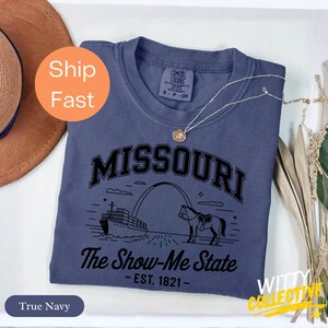 Puede incluir: Una camiseta azul marino con la palabra "MISSOURI" sobre una ilustraci&oacute;n de un barco, un arco y un caballo. El texto "The Show-Me State - EST. 1821-" est&aacute; debajo. Un collar dorado est&aacute; sobre la camiseta.