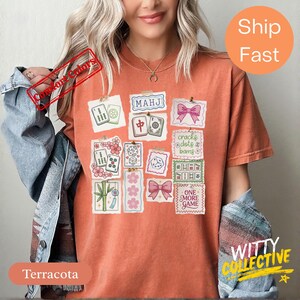 May include: Terracotta-colored t-shirt featuring a collage of Mahjong tile illustrations, pink bows, and floral accents. The shirt includes the text "MAHJ" and "ONE MORE GAME".
