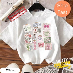 May include: White t-shirt featuring a collage of Mahjong tile illustrations, pink bows, and the text "MAHJ". The shirt includes the phrases "cracks dots bams" and "ONE MORE GAME". The shirt has short sleeves and a round neckline.