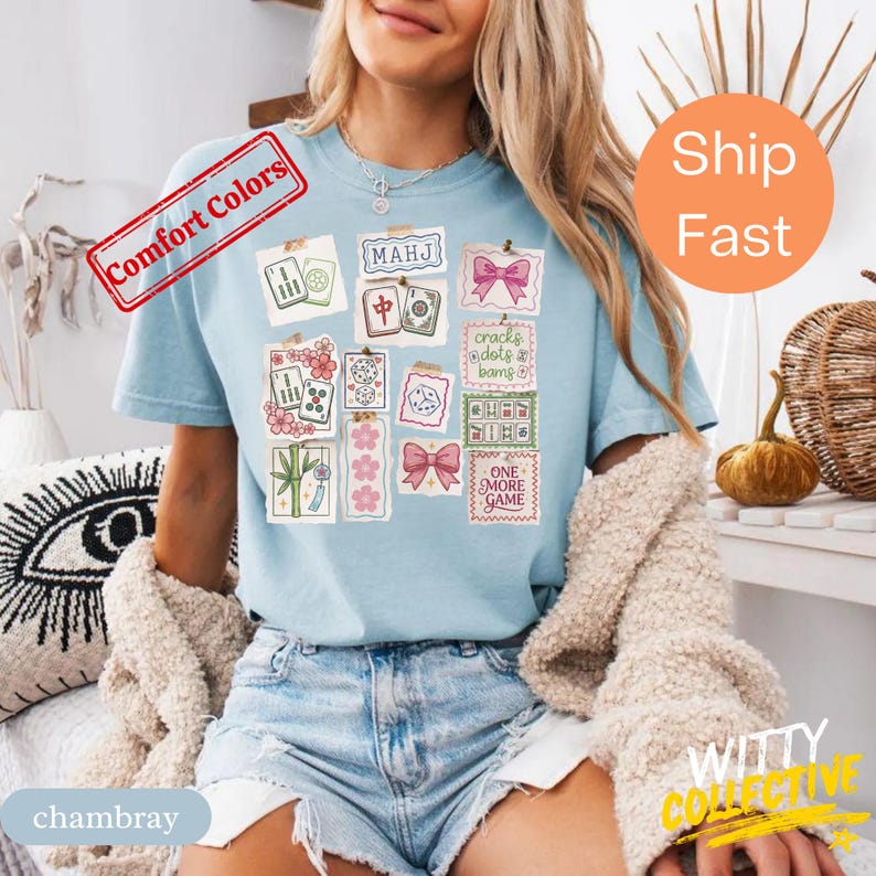May include: Light blue t-shirt featuring a collage of Mahjong tile illustrations and decorative elements. The design includes text that reads "MAHJ", "cracks dots & bams", and "ONE MORE GAME". The shirt has short sleeves and a crew neck.