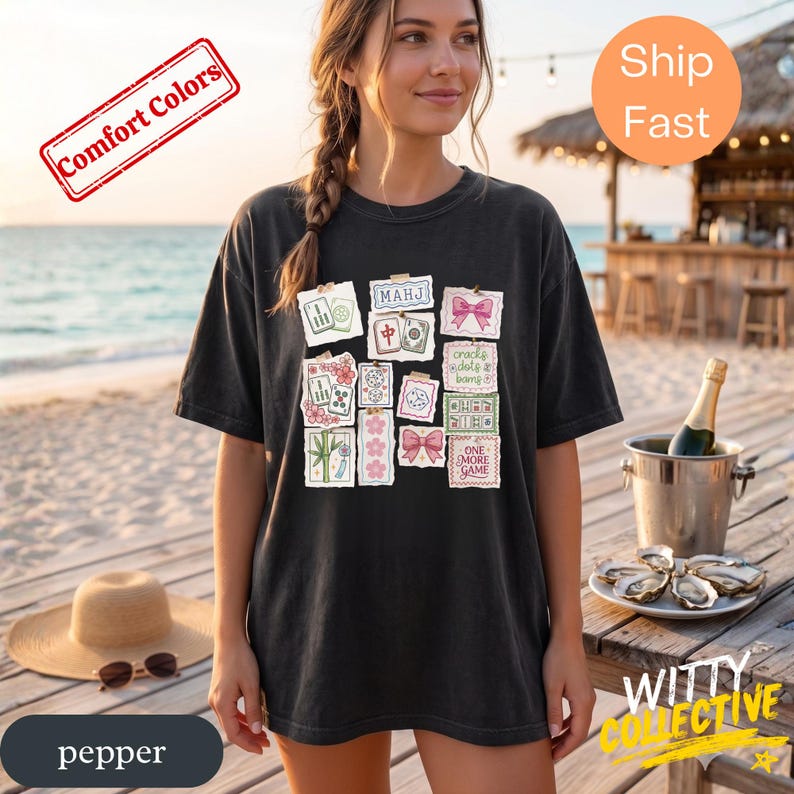 May include: Black t-shirt with a graphic of Mahjong tiles and pink bows. The shirt has the text "MAHJ" and "ONE MORE GAME". The shirt is a comfort color and is pepper colored.