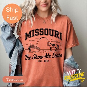 Puede incluir: Camiseta de color terracota con la palabra "MISSOURI" sobre una ilustraci&oacute;n de un barco, un arco y un caballo. El texto "The Show-Me State - EST. 1821-" est&aacute; debajo. La camiseta tiene mangas cortas.