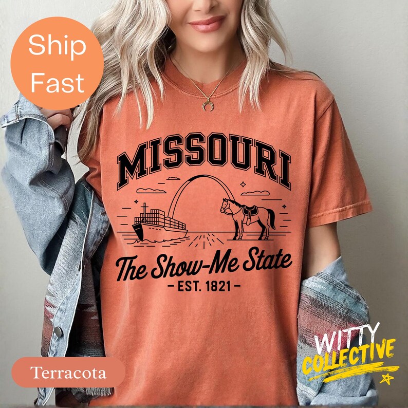 Puede incluir: Camiseta de color terracota con la palabra "MISSOURI" sobre una ilustraci&oacute;n de un barco, un arco y un caballo. El texto "The Show-Me State - EST. 1821-" est&aacute; debajo. La camiseta tiene mangas cortas.
