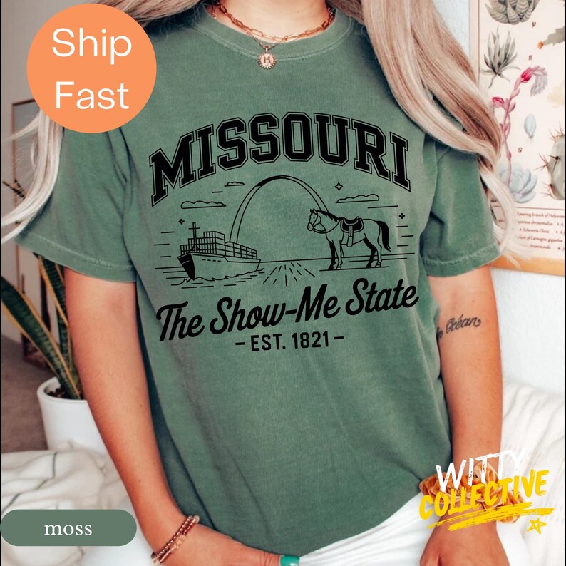 Puede incluir: Una camiseta verde musgo con la palabra "MISSOURI" en negro sobre un gr&aacute;fico del Gateway Arch, un caballo y un barco. El texto "The Show-Me State - EST. 1821-" est&aacute; debajo.