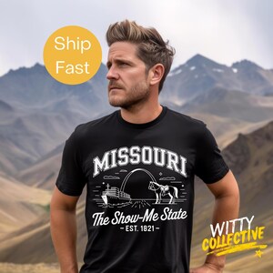 Puede incluir: Camiseta negra con un dise&ntilde;o gr&aacute;fico blanco que presenta la palabra "MISSOURI" sobre una ilustraci&oacute;n de un barco, un caballo y un arco. El texto "The Show-Me State - EST. 1821 -" est&aacute; debajo. La camiseta la lleva una persona.