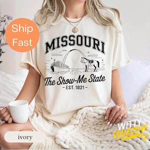 Puede incluir: Camiseta de color marfil con un dise&ntilde;o gr&aacute;fico negro. El dise&ntilde;o incluye la palabra "MISSOURI", una ilustraci&oacute;n del Gateway Arch, un barco y un caballo. El texto "The Show-Me State - EST. 1821-" est&aacute; debajo.