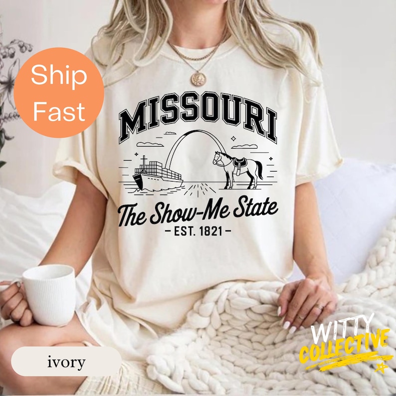 Puede incluir: Camiseta de color marfil con un dise&ntilde;o gr&aacute;fico negro. El dise&ntilde;o incluye la palabra "MISSOURI", una ilustraci&oacute;n del Gateway Arch, un barco y un caballo. El texto "The Show-Me State - EST. 1821-" est&aacute; debajo.