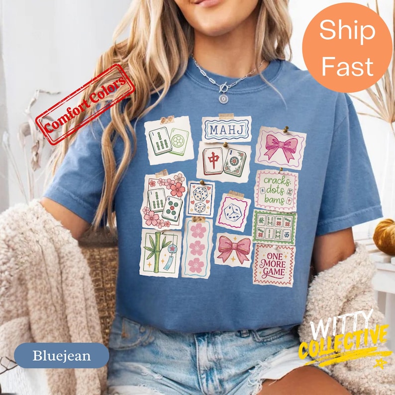 May include: Blue jean colored t-shirt with a collage of Mahjong tile illustrations. The design includes text that reads "MAHJ", "cracks dots bams", and "ONE MORE GAME". The shirt has a relaxed fit and is made of a soft material.