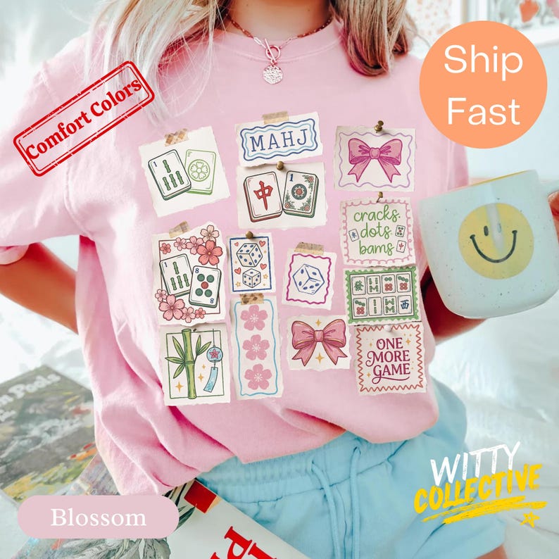 May include: A light pink sweatshirt with a collage of Mahjong tiles, dice, and floral designs. The shirt features the text "MAHJ" and "ONE MORE GAME". A white mug with a yellow smiley face is held in the hand.