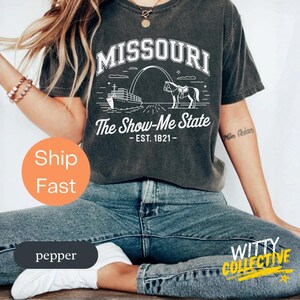 Puede incluir: Camiseta gris oscuro con un gr&aacute;fico blanco del arco de Missouri, un barco y un caballo. El texto dice "MISSOURI" arriba y "The Show-Me State - EST. 1821-" abajo. La camiseta es de color pimienta.